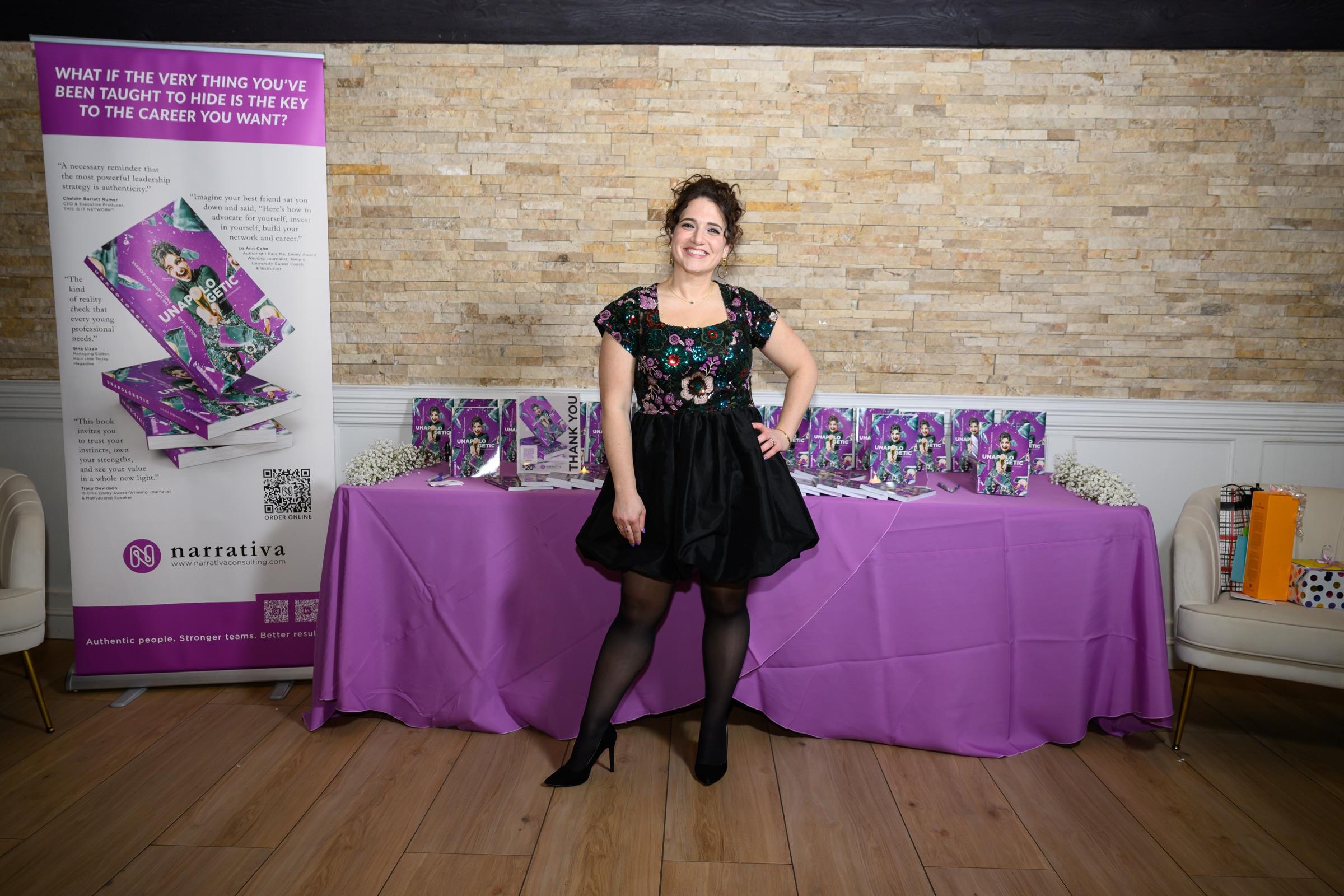 Nicole at her Unapologetic book launch, beside a purple-draped table of copies.